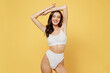 © ViDi Studio - Smiling slim confident beautiful lovely attractive young brunette woman 20s in white underwear with perfect fit body standing posing raise up hands isolated on plain yellow background studio portrait.
