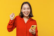 Jubilant vivid excited fancy young woman of Asian ethnicity 20s years old wear orange shirt hold in hand use mobile cell phone doing winner gesture isolated on plain yellow background studio portrait