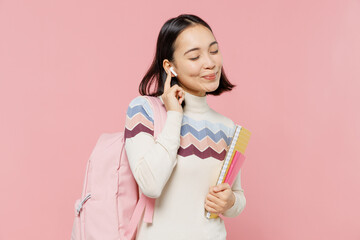 Wall Mural - Satisfied happy teen student girl of Asian ethnicity wear sweater backpack hold books listen music in earphones isolated on pastel plain light pink background Education in university college concept