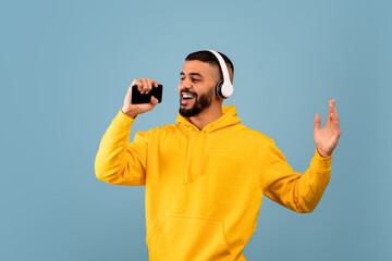 Young arab man singing, holding smartphone like microphone wearing wireless headphones and having fun, blue background