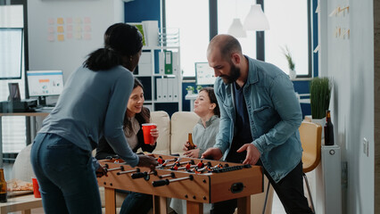 Wall Mural - Diverse group of workmates enjoying game with foosball table to do fun activity after work. Coworkers playing football and drinking beer from bottles, celebrating party with entertainment