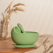 Modern green silicone bowl with suction base and spoon on wooden table near glass vase with dried flowers. Serving food, baby tableware, first feeding concept. Instagram use, square frame, soft focus.