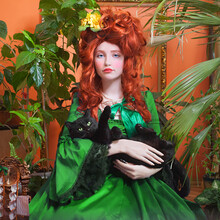 Portrait Of A Girl With A Beautiful Hairstyle In The Rococo Style In A Green Dress Against Plants' Background. Red-haired Young Woman With Black Cat. Indoors Shot In The Marie Antoinette Style.
