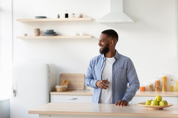 Cheerful young bearded black guy with glass of clean water in hand in kitchen interior looks at window