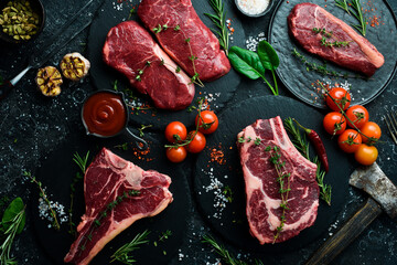 A variety of raw beef steaks for grilling with seasonings: T-bone, striploin steak, ribeye. Top view. On a black stone background.