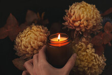 Hand Holding A Scented Candle In A Brown Glass Jar Near Flowers Bouquet, Aromatherapy And Spa. Glowing Burning Candle And Flowers Brown Background. Spa, Aromatherapy And Treat Yourself Concept.