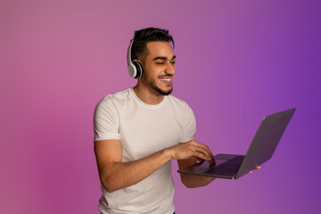 Poster - Positive millennial Arab man in wireless headphones using laptop pc for online work or communication in neon light