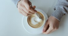 female hands in a gray sweater beautifully and slowly stir a cappuccino with foam in a white cup on a wooden table with a spoon. coffee tasting. view from above