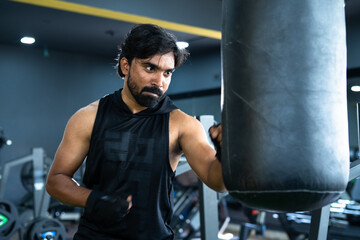 Angry young Indian athlete punching boxing bag at gym - concept of motivational, hardworking, fighting and workout training