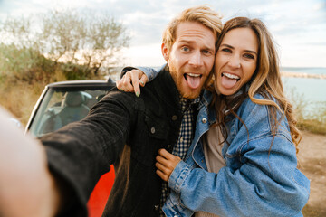 Wall Mural - White couple making fun while taking selfie photo by car during trip