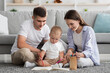 © Prostock-studio - Leisure With Baby. Young Happy Parents And Toddler Child Drawing At Home