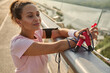 Confident relaxed middle aged sportswoman with a bottle of water and a skipping rope, looking away while admiring beautiful nature standing on the city bridge after intense cardio workout at sunrise