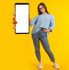 Poster - Mobile app recommendation. Big smartphone in woman hand. Girl in full growth with smartphone. Phone with blank white screen. Mock up phone for your advertisement. Mock up phone on yellow background