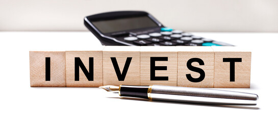 Fototapete - The light table has a black calculator, a fountain pen, and wooden cubes with the text INVEST. Business concept