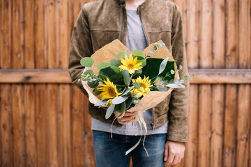 Very nice young man holding big and beautiful mono bouquet of fresh yellow sunflowers, cropped photo, bouquet close up on the wooden background