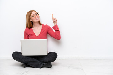 Wall Mural - Young redhead woman with a laptop sitting on the floor isolated on white background showing and lifting a finger in sign of the best