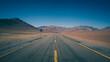 Eine asphaltierte Straße führt durch die Atacama Wüste Chiles entlang roter Sandlandschaften sowie riesiger Vulkane und schneebedeckter Berge