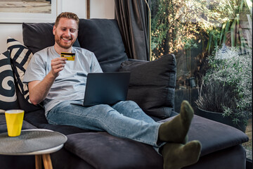 Wall Mural - Happy cheerful smiling young adult man doing online shopping or e-shopping satisfied entrepreneur making online payment paying for service or goods while comfortably sitting on sofa at his home