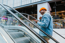 Joyful Young African-American Man In Stylish Clothes Looks At Mobile Phone Screen Going Up Escalator In Large Shopping Mall