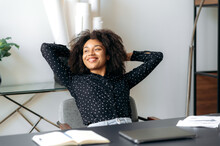 African American Satisfied Pretty Girl, Office Worker, Manager, Sits At Work Desk, Takes A Break From Work, Looks To The Side, Throwing Her Hands Behind Her Head, Smiling, Dreaming About Vacation