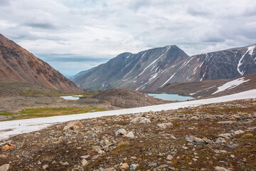 Dramatic landscape with glacier and stone hill between two mountain lakes in sunlight under cloudy sky. Two small lake in high mountain valley with view to mountain vastness at changeable weather.