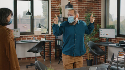 Happy employee dancing after winning successful deal, wearing face mask. Business man celebrating achievement and feeling excited while woman looking at playful colleague. Funny worker