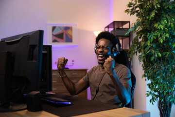 Winning a round in a computer video game. A dark-skinned man with a headset shouts to the team for the joy of victory, a gamer clenches fists in triumph, the room dark with colored led lights