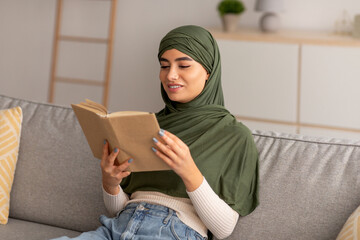 Wall Mural - Peaceful domestic hobbies. Cheery young Arab woman in hijab reading book, relaxing on sofa in living room