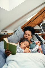 Wall Mural - Small African American girl fell asleep while mother is reading her book on the sofa.