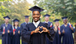 © Syda Productions - education, graduation and people concept - happy graduate student woman in mortarboard and bachelor gown showing hand heart gesture over group of people on background
