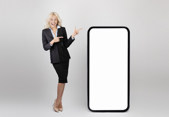 Canvas Print - Business advertisement. Young happy businesswoman pointing at big smartphone with white screen, mockup