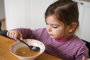 Close up of preschool girl portrait with funny expression eating soup at home 