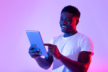 Portrait of joyful young black guy using tablet pc, studying or working remotely, having online meeting in neon light