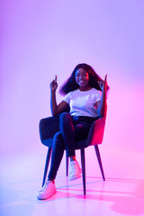 Wall Mural - Smiling young black woman sitting in armchair, pointing upwards, offering blank space in neon light, mockup for ad