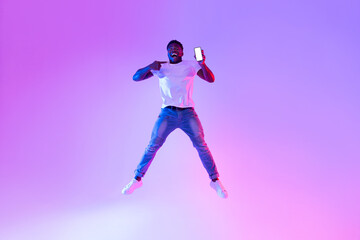 Poster - Full length of excited young black guy jumping with cellphone, pointing at empty screen in neon light, mockup