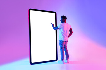 Poster - Happy young African American man standing near big tablet computer with mockup, touching screen in neon light