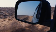 View Of Side Mirror Of Car, Which Reflects Unpaved Road Behind Vehicle, Dry Plants, Sky, Electric Power Transmission Poles. Sunny Summer Day In Barren Area. Auto Driving On Soil Route In Desert Place
