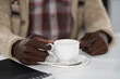 © Svyatoslav Lypynskyy - Hands of African American man holding cup of coffee wearing beige sweater sitting at the table at the campus cafeteria. Drops of coffee on spoon isolated on white table