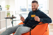 © Prostock-studio - Cheerful Man Shopping Using Phone And Credit Card Sitting Indoors