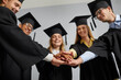 © Studio Romantic - Close up of excited college graduates in mantles and hats join stack hands in pile show unity and leadership. Smiling young students in robes celebrate university graduation. Education concept.