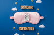 Top view photo of one big pink blindfold and small white alarm on it with scattered confetti in shape of stars and clouds and wooden cubes with words on the deep blue background