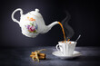 Levitating teapot pouring tea into cup with steam, cinnamon tied with rope, on gray table on dark background, white porcelain service