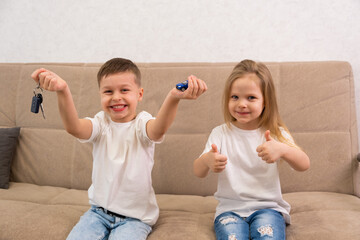 A happy boy in a white T-shirt holds keys and a toy car in his hands and looks at the camera. The girl gives a thumbs up. The concept of buying and winning a family car