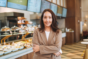 Adult smiling brunette business woman with long hair in stylish beige suit with mobile phone looking at camera owner of small business cafe bakery