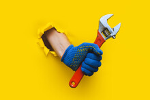 A Left Hand In A Blue Knitted Glove Holds A Red Monkey Wrench. Torn Hole In Yellow Paper. The Concept Of A Worker, Labor Migrant, A Master Of His Craft. Copy Space.