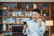 Close-up portrait of a successful male businessman working in a classic office, Asian won, in the online casino app, looks at the phone screen and rejoices in victory