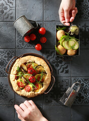 Wall Mural - Homemade cakes. Galette with vegetables: potatoes, eggplant, zucchini, cherry tomatoes and arugula with hands on a gray kitchen table. Dough with vegetables and spices on gray plate.Top view, flatlay