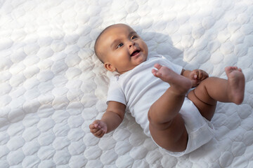 Happy dark skinned baby 5 months old baby lies in the nursery white clothes on her back and smile, looks at someone,  black infant child lies on selective focus, with copy space