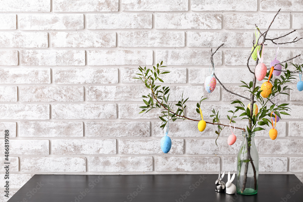 Green tree branches with Easter eggs and rabbits on table near light brick wall
