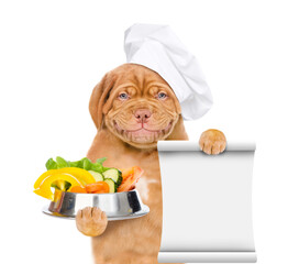 Poster - Smiling puppy wearing a chef's cap  holds bowl of vegetables and empty list. isolated on white background